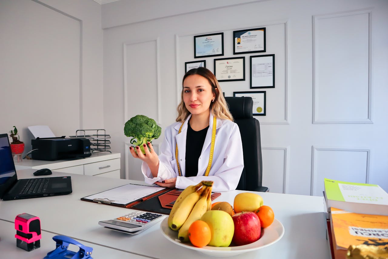 service-03 Female nutritionist in office holding broccoli, surrounded by fruits, promoting healthy lifestyle.